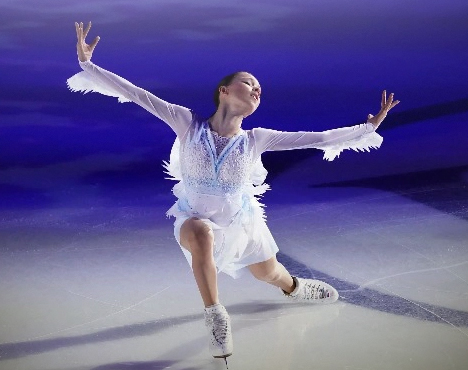 Фигуристка в белом костюме выполняет эффектный элемент на льду. Она стоит на одном колене, руки вытянуты в стороны. Фон сияет синими и фиолетовыми оттенками, создавая атмосферу драмы и грации