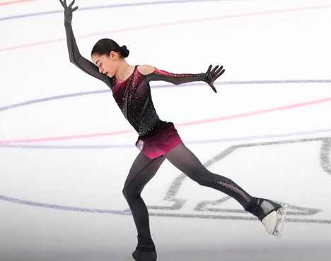 Фигуристка в костюме градиентных оттенков черного и бордового выполняет элемент на льду. На заднем плане видна разметка катка