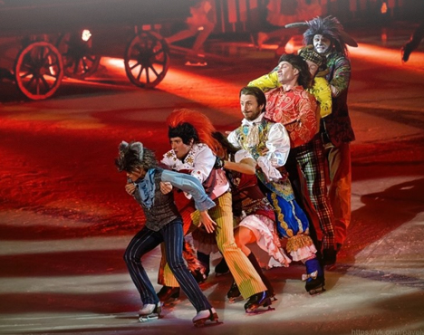 На ледяной арене группа людей в ярких, разнообразных костюмах катается на коньках. Они выстроились друг за другом, создавая впечатление веселого шествия. Атмосфера мероприятия праздничная и динамичная